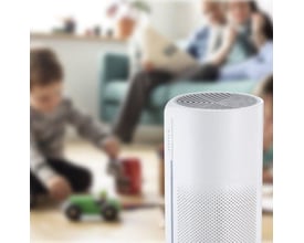Sensibo Pure luftrenser i hvid står på et gulvtæppe, mens en familie leger og slapper af i baggrunden.