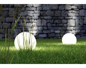 TRIO Lighting Bolo Spjut gulvlampe i sort plast, oplyst og placeret i græsset foran en stenmur.