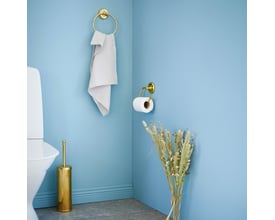 Smedbo Villa toiletpapirholder i poleret messing monteret på lyseblå væg ved siden af et toilet og et håndklædestativ.