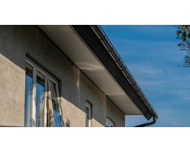 Plastmo sort tagrende monteret på en lys facade med hvide vinduer mod en klar blå himmel.