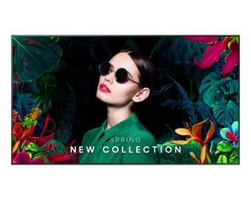 Samsung QM98C-skærm med sort ramme viser en kvinde foran farverige tropiske planter og teksten »New Collection«.