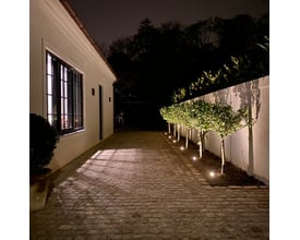 Light By Juul chaussesten lyser en brostensbelagt indkørsel og op ad en mur med effektfuldt lys på træer og buske.
