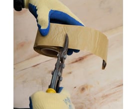 En hånd med gul og blå arbejdshandske holder en rulle NI Metex rottetape og anvender en papegøjetang til at klippe tapen af, klar til reparation af træoverflader.