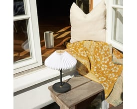 Halo Design Paris to go sort metal bordlampe med plisseret skærm placeret på et rustikt træbord med guldfarvet tæppe og pude.