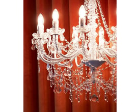 Nærbillede af den 8-armede Nielsen Light Imperial lysekrone med hvidt stel og klare glasprismer mod en orange baggrund.