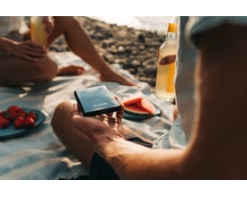 En person holder en sort Verbatim Store 'n' Go Slim Portable SSD på en strand med jordbær og drikkevarer i baggrunden.