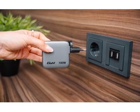 Verbatim mini GaN-oplader på 100 W med display holdes i hånden ved siden af en stikkontakt.