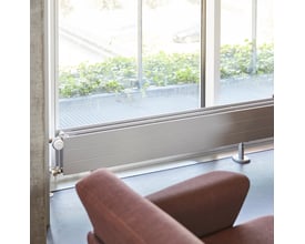 Hudevad LK konvektor model T34/3-30 i RAL9016 hvid, monteret ved et stort vindue med udsigt til grønne planter.