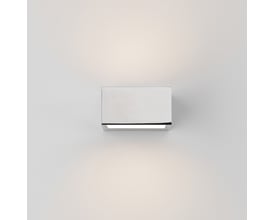 Astro 8162 Kappa LED væglampe i blank krom, tændt med varmt hvidt lys på hvid væg.