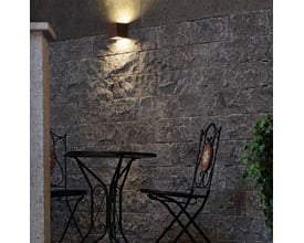 SLV Sitra væglampe i rustfarvet aluminium op/ned, monteret på en rå murstensvæg over et havebord og stole.