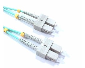 Fiberoptisk patchkabel med to SC/PC konnektorer i grå plast, med lyseblå kabel og gule manchetter, på hvid baggrund.