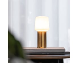 SACKit Base London lampefod i messing med tændt, hvid lampeskærm, placeret på en bog på et bord.