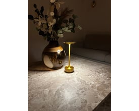 Elegant Skagen genopladelig LED lampe i guld lyser op på et marmorbord ved siden af en vase med blomster og grønne planter.