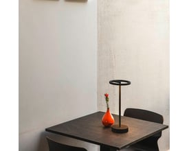 SLV VINOLINA ONE akku bordlampe i sort aluminium står på et mørkt bord ved siden af en orange vase med en rød blomst.