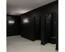 SLV One Linear væglampe i mat sort, 70 cm lang, diskret monteret på en mørk væg i et moderne rum med lysegrå gulv.