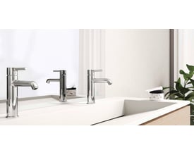 To Oras Optima Style krom håndvaskarmaturer står på en hvid vask, med et spejl og en grøn plante i baggrunden.