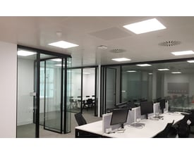 SG Armaturen Sense Pro X 600x600mm panel med 4000K hvidt lys, installeret i et moderne kontor med glasvægge og skriveborde.