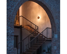 SLV Mana Base Phase væglampe i antracit grå monteret på en væg ved en trappe, lyser varmt.