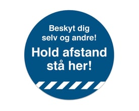 Blåt rundt gulvskilt med hvid tekst "Beskyt dig selv og andre! Hold afstand - stå her!" og hvide skråstriber nederst.
