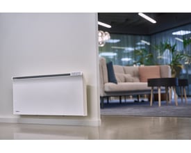 Glamox Termostat WT3 WiFi monteret på væg i et moderne kontormiljø med sofa og planter i baggrunden.