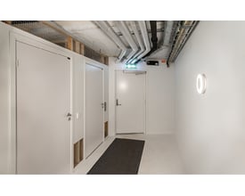 SG Armaturen Etne væg-/loftarmatur i hvidt polycarbonat med 4000K lys på en hvid væg i en gang med døre og rør i loftet.