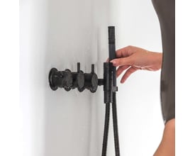 Vola indbygningstermostat i matsort med tre greb og håndbruser, der betjenes af en hånd på en hvid flisevæg med vandsprøjt.