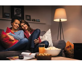 LEDVANCE HS mat LED-kronepære med E14-fatning lyser varmt i en gulvlampe ved siden af en sofa.