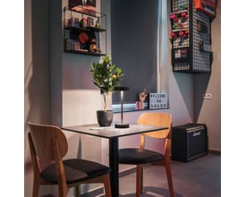 SLV VINOLINA bordlampe i sort aluminium, tændt og placeret på et bord med to stole og en guitarforstærker i baggrunden.