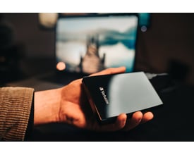 Verbatim Store ´n´ Go Slim Portable SSD i sort holdes i en hånd med en bærbar computer i baggrunden.