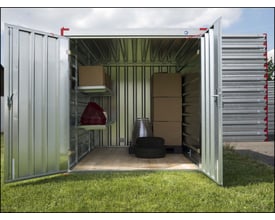 Åbent opbevaringscontainer med metalhylder, der indeholder papkasser, en rød trekant advarsel og to dæk. Containeren står på en græsplæne.