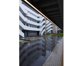 Moderne bygning med udendørs belysning, der reflekterer på våde fliser og asfaltsti.