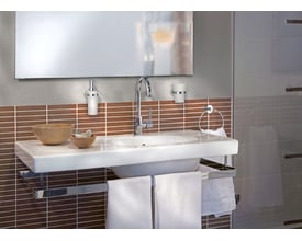 Smedbo home sæbedispenser i messing og materet glas monteret på væggen ved siden af et badeværelsesvask og spejl.