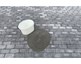En hvid spand og en bunke A-JOINT Polysand PRO specialfugesand i stone grey placeret på en grå stenbelægning.