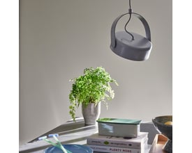 Detaljebillede af den grå Hübsch Stage loftlampe, der hænger over en potteplante og bøger på en bordplade.