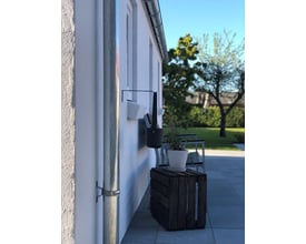 Plastmo tudstykke i zink monteret på nedløbsrør ved en hvid facade, med have og træ i baggrunden.