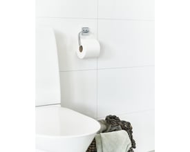 Smedbo toiletpapirholder i poleret krom monteret på et hvidt, flisebelagt badeværelsesvæg med en toiletpapirrulle påsat.