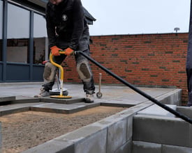 Person med Ravendo Mini Vakuum TL fliseløfteren placeret på fliser og tilsluttet en støvsuger, mens der arbejdes udendørs.