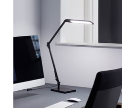 Nielsen Light arkitektlampe i sort, med justerbar arm og lampehoved der lyser, placeret på et kontorbord ved siden af en computerskærm.