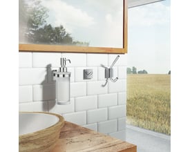 Smedbo House sæbedispenser i materet glas med poleret krom pumpe, monteret på en hvid flisevæg ved siden af et håndvaskområde.