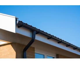 Plastmo ekspansionsstykke nr. 10 monteret på et mørkegråt tagrendesystem med nedløbsrør på en husmur under en klar blå himmel.