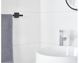 Smedbo House håndklædestang i mat sort messing, monteret på en hvid flisevæg ved siden af en hvid vask og krom vandhane.