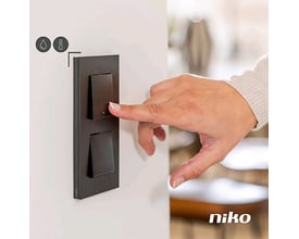 Niko 2-tryk til Niko Home Control i sort med LED og komfortsensorer, vist med en finger der trykker på knappen.