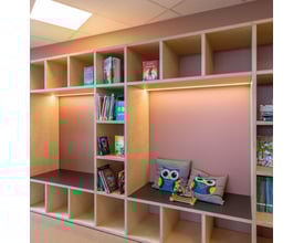 Indbygget reol med lyserøde vægge og åbne rum fyldt med bøger, legetøj og dekorative puder med uglemotiv.