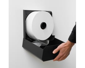Nichba jumbo papirholder ses monteret med en stor toiletrulle, mens en hånd trækker papir ud.