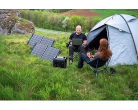 100W solpanel sat op i naturen ved siden af et telt, med to personer der sidder i stole og bruger en bærbar computer, mens strøm fra solpanelet oplader udstyr.