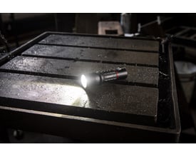 Coast XP11R lygte med lysende LED-pære, placeret på et rustent metalbord i et værkstedsmiljø.