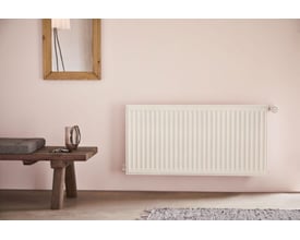 Stelrad Compact All In Type 21 hvid radiator monteret på lyserød væg med et træbænk og spejl i rummet.