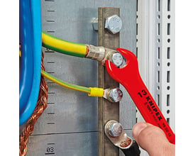 Hånd der bruger en rød KNIPEX L-AUS fastnøgle til at spænde en bolt på en elektrisk samleskinne med flere kabler tilsluttet.