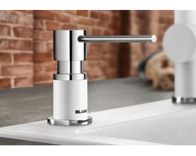 BLANCO LATO sæbedispenser i klippegrå SILGRANIT® og skinnende krom, monteret ved siden af en hvid køkkenvask med murstensvæg i baggrunden.