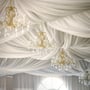 Elegant gyldent lysekrone med krystalvedhæng hænger fra et hvidt draperet loft i festlig atmosfære.